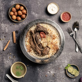 A plate of flavorful biryani garnished with roasted meat, a whole garlic bulb, and a charred tomato sits on a rustic background. Surrounding the main dish are small bowls containing condiments like yogurt, red sauce, spicy green soup, and gulab jamun. A sprig of mint and lime slices are arranged nearby, with cutlery placed neatly to the side.
