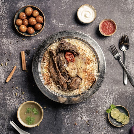 A plate of flavorful biryani garnished with roasted meat, a whole garlic bulb, and a charred tomato sits on a rustic background. Surrounding the main dish are small bowls containing condiments like yogurt, red sauce, spicy green soup, and gulab jamun. A sprig of mint and lime slices are arranged nearby, with cutlery placed neatly to the side.