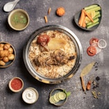 A gourmet dish featuring a large metal bowl filled with biryani, topped with a cooked chicken leg, roasted tomato, and slices of onion, accompanied by a variety of sides including a soup with cilantro garnish, small roasted potatoes, sliced cucumbers and carrots, sliced tomatoes and onions, a bowl of red sauce, a bowl of yogurt, and sliced limes with mint leaves. There are also assorted spices and a decorative piece resembling a knife on the side.