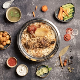A gourmet dish featuring a large metal bowl filled with biryani, topped with a cooked chicken leg, roasted tomato, and slices of onion, accompanied by a variety of sides including a soup with cilantro garnish, small roasted potatoes, sliced cucumbers and carrots, sliced tomatoes and onions, a bowl of red sauce, a bowl of yogurt, and sliced limes with mint leaves. There are also assorted spices and a decorative piece resembling a knife on the side.