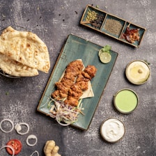 A overhead view of a food spread featuring grilled chicken pieces placed on a rectangular blue plate, accompanied by sliced onions, a lime wedge, and some herbs. Surrounding the plate are bowls containing sauces of various colors, a stack of naan bread, and an assortment of spices in a dish. The textured stone-like background adds to the rustic aesthetic.
