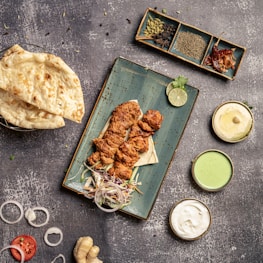 A overhead view of a food spread featuring grilled chicken pieces placed on a rectangular blue plate, accompanied by sliced onions, a lime wedge, and some herbs. Surrounding the plate are bowls containing sauces of various colors, a stack of naan bread, and an assortment of spices in a dish. The textured stone-like background adds to the rustic aesthetic.