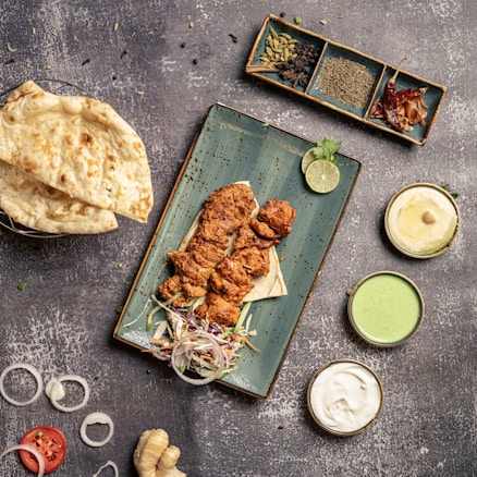 A overhead view of a food spread featuring grilled chicken pieces placed on a rectangular blue plate, accompanied by sliced onions, a lime wedge, and some herbs. Surrounding the plate are bowls containing sauces of various colors, a stack of naan bread, and an assortment of spices in a dish. The textured stone-like background adds to the rustic aesthetic.