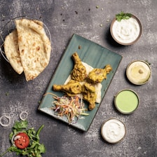 A table is set with a meal featuring a rectangular green plate with three pieces of grilled or tandoori chicken on naan bread, accompanied by a colorful coleslaw. To the side, there are three sauces in small bowls: one white, one light green, and one beige. A basket contains two pieces of flatbread, likely naan, and there is a garnish of fresh cilantro and a sliced tomato with onion rings.