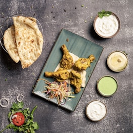 A table is set with a meal featuring a rectangular green plate with three pieces of grilled or tandoori chicken on naan bread, accompanied by a colorful coleslaw. To the side, there are three sauces in small bowls: one white, one light green, and one beige. A basket contains two pieces of flatbread, likely naan, and there is a garnish of fresh cilantro and a sliced tomato with onion rings.