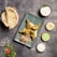 A table is set with a meal featuring a rectangular green plate with three pieces of grilled or tandoori chicken on naan bread, accompanied by a colorful coleslaw. To the side, there are three sauces in small bowls: one white, one light green, and one beige. A basket contains two pieces of flatbread, likely naan, and there is a garnish of fresh cilantro and a sliced tomato with onion rings.