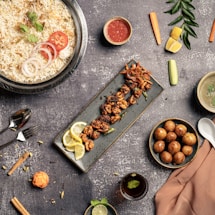 An assortment of delicious dishes is arranged artfully on a textured gray tabletop. A large bowl of rice is garnished with coriander, tomato, and onion slices. Next to it, a long rectangular plate holds skewered grilled meat garnished with lemon slices. A small bowl of red sauce, a bowl of soup, a spoon, and a fork are placed nearby. On the right side, a bowl filled with round golden-brown appetizers is visible. Vegetables such as carrots, cucumbers, and lemon wedges are scattered around, adding pops of color alongside a small glass of beverage with mint.