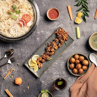 An assortment of delicious dishes is arranged artfully on a textured gray tabletop. A large bowl of rice is garnished with coriander, tomato, and onion slices. Next to it, a long rectangular plate holds skewered grilled meat garnished with lemon slices. A small bowl of red sauce, a bowl of soup, a spoon, and a fork are placed nearby. On the right side, a bowl filled with round golden-brown appetizers is visible. Vegetables such as carrots, cucumbers, and lemon wedges are scattered around, adding pops of color alongside a small glass of beverage with mint.