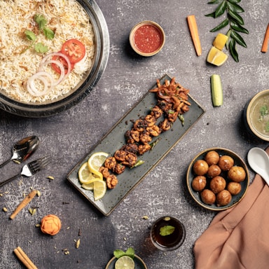 An assortment of delicious dishes is arranged artfully on a textured gray tabletop. A large bowl of rice is garnished with coriander, tomato, and onion slices. Next to it, a long rectangular plate holds skewered grilled meat garnished with lemon slices. A small bowl of red sauce, a bowl of soup, a spoon, and a fork are placed nearby. On the right side, a bowl filled with round golden-brown appetizers is visible. Vegetables such as carrots, cucumbers, and lemon wedges are scattered around, adding pops of color alongside a small glass of beverage with mint.
