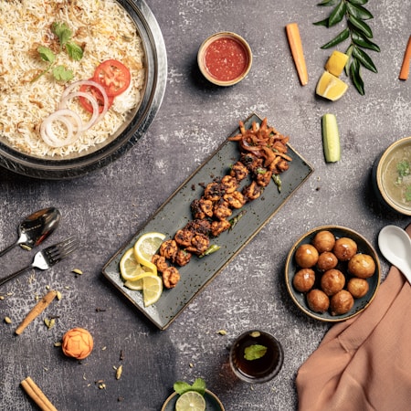 An assortment of delicious dishes is arranged artfully on a textured gray tabletop. A large bowl of rice is garnished with coriander, tomato, and onion slices. Next to it, a long rectangular plate holds skewered grilled meat garnished with lemon slices. A small bowl of red sauce, a bowl of soup, a spoon, and a fork are placed nearby. On the right side, a bowl filled with round golden-brown appetizers is visible. Vegetables such as carrots, cucumbers, and lemon wedges are scattered around, adding pops of color alongside a small glass of beverage with mint.