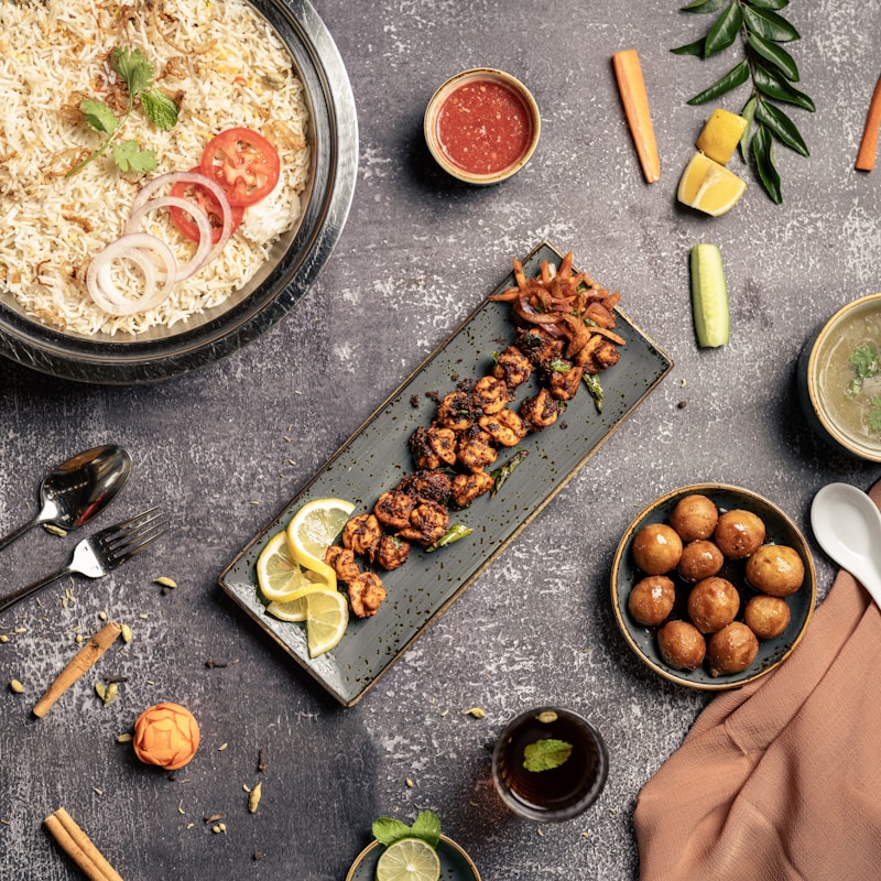 An assortment of delicious dishes is arranged artfully on a textured gray tabletop. A large bowl of rice is garnished with coriander, tomato, and onion slices. Next to it, a long rectangular plate holds skewered grilled meat garnished with lemon slices. A small bowl of red sauce, a bowl of soup, a spoon, and a fork are placed nearby. On the right side, a bowl filled with round golden-brown appetizers is visible. Vegetables such as carrots, cucumbers, and lemon wedges are scattered around, adding pops of color alongside a small glass of beverage with mint.