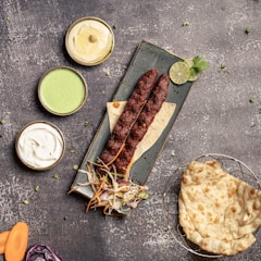 A rectangular plate with two skewered kebabs garnished with sliced lime and cilantro is placed on a table. Next to it are bowls containing hummus, mint chutney, and yogurt. Accompanying them are slices of flatbread and a small salad of shredded carrots and cabbage. The setting is casual, with ingredients artistically arranged.