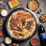 A large dish containing spiced rice with various types of meat, including grilled chicken and lamb, served on a round tray. Surrounding the main dish are sides such as yogurt, sliced vegetables, sauce, a bottle of Pepsi, and different small bowls containing sweets or condiments. The background is a dark, textured surface with scattered spices and herbs.