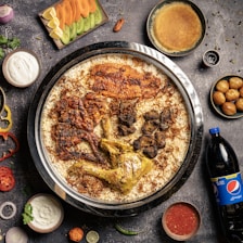 A large dish containing spiced rice with various types of meat, including grilled chicken and lamb, served on a round tray. Surrounding the main dish are sides such as yogurt, sliced vegetables, sauce, a bottle of Pepsi, and different small bowls containing sweets or condiments. The background is a dark, textured surface with scattered spices and herbs.