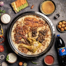 A large dish containing spiced rice with various types of meat, including grilled chicken and lamb, served on a round tray. Surrounding the main dish are sides such as yogurt, sliced vegetables, sauce, a bottle of Pepsi, and different small bowls containing sweets or condiments. The background is a dark, textured surface with scattered spices and herbs.