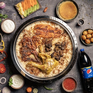A large dish containing spiced rice with various types of meat, including grilled chicken and lamb, served on a round tray. Surrounding the main dish are sides such as yogurt, sliced vegetables, sauce, a bottle of Pepsi, and different small bowls containing sweets or condiments. The background is a dark, textured surface with scattered spices and herbs.