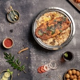 A top-down view of a table with food items including a large plate of rice topped with a piece of grilled fish garnished with herbs. Surrounding the main dish are various smaller items: a small bowl of red sauce, a sprig of green leaves, sliced lime, onion rings, tomato slices, a dark beverage with a mint leaf, a small bowl of golden brown balls, and some golden measuring spoons on a plate.