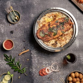 A top-down view of a table with food items including a large plate of rice topped with a piece of grilled fish garnished with herbs. Surrounding the main dish are various smaller items: a small bowl of red sauce, a sprig of green leaves, sliced lime, onion rings, tomato slices, a dark beverage with a mint leaf, a small bowl of golden brown balls, and some golden measuring spoons on a plate.