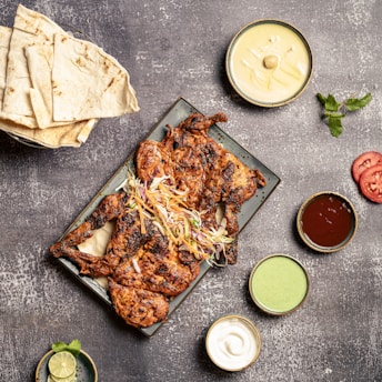 A grilled, spiced chicken dish served on a rectangular plate garnished with shredded vegetables. Surrounding the plate are various small bowls containing dipping sauces, and some tomato slices. To the left, there is a stack of flatbread, and a small bowl with lime and mint.