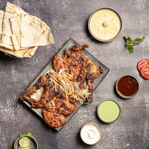A grilled, spiced chicken dish served on a rectangular plate garnished with shredded vegetables. Surrounding the plate are various small bowls containing dipping sauces, and some tomato slices. To the left, there is a stack of flatbread, and a small bowl with lime and mint.