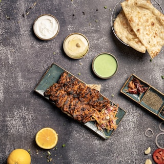 A platter of grilled, marinated meat skewers served with a colorful slaw, surrounded by small bowls of sauces including white, beige, and green dips. Two pieces of flatbread are placed in a basket next to the dips. A halved lemon, spices, and a variety of garnishes such as sliced onion and peppers are scattered on the textured surface.