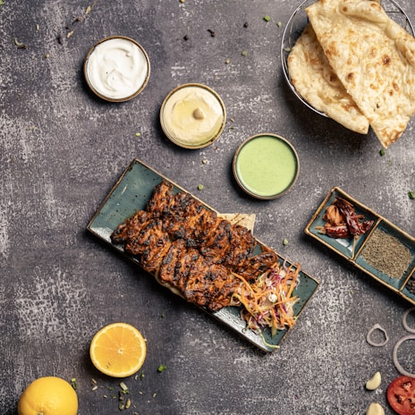 A platter of grilled, marinated meat skewers served with a colorful slaw, surrounded by small bowls of sauces including white, beige, and green dips. Two pieces of flatbread are placed in a basket next to the dips. A halved lemon, spices, and a variety of garnishes such as sliced onion and peppers are scattered on the textured surface.