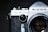 A close-up view of an old-fashioned camera with a textured black grip and a silver top. The name 'Ricoh' is prominent on the top panel, and a dial indicating 'ASA' is visible. The lens is partially in the frame with reflections on its glass surface.