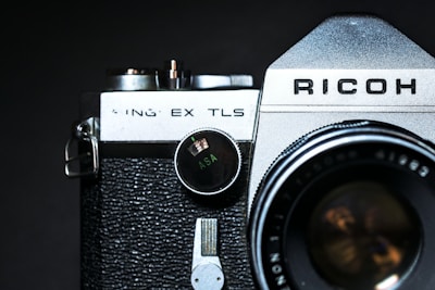 A close-up view of an old-fashioned camera with a textured black grip and a silver top. The name 'Ricoh' is prominent on the top panel, and a dial indicating 'ASA' is visible. The lens is partially in the frame with reflections on its glass surface.
