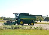 A green agricultural combine harvester with yellow accents is parked on a farm field. The background includes open fields and sparse trees under a clear sky.
