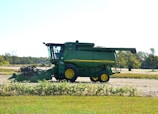 A green agricultural combine harvester with yellow accents is parked on a farm field. The background includes open fields and sparse trees under a clear sky.