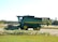A green agricultural combine harvester with yellow accents is parked on a farm field. The background includes open fields and sparse trees under a clear sky.