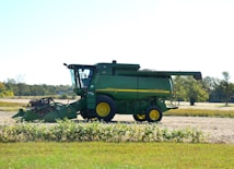 A green agricultural combine harvester with yellow accents is parked on a farm field. The background includes open fields and sparse trees under a clear sky.