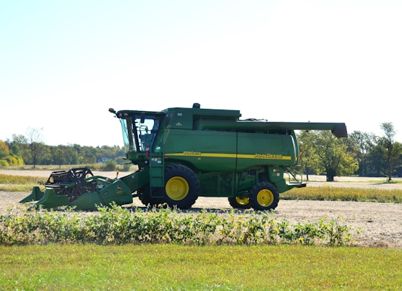 A green agricultural combine harvester with yellow accents is parked on a farm field. The background includes open fields and sparse trees under a clear sky.