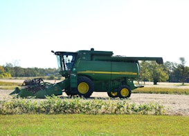 A green agricultural combine harvester with yellow accents is parked on a farm field. The background includes open fields and sparse trees under a clear sky.