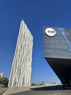A modern, angular white building with a crisscross pattern stands against a clear blue sky, alongside a dark, geometric structure featuring a sign with the text 'nat' in bold letters.