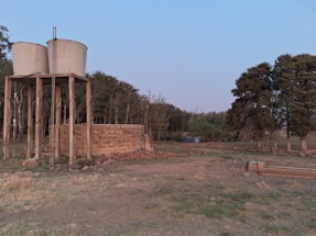 Two cylindrical water tanks are elevated on a wooden structure next to a stone wall. The foreground features dry, grassy terrain with scattered rocks. In the background, there is a line of dense trees with a small pond visible through them. The sky is clear with a subtle gradient.
