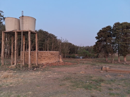 Two cylindrical water tanks are elevated on a wooden structure next to a stone wall. The foreground features dry, grassy terrain with scattered rocks. In the background, there is a line of dense trees with a small pond visible through them. The sky is clear with a subtle gradient.
