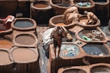 A traditional tannery with several circular stone pits filled with colored liquid can be seen. Workers are engaged in their tasks, handling pieces of leather and fabric. The scene reflects the labor-intensive process of leather tanning, with the earthy tones of the stone contrasting with the vibrant hues of the dye.