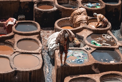 A traditional tannery with several circular stone pits filled with colored liquid can be seen. Workers are engaged in their tasks, handling pieces of leather and fabric. The scene reflects the labor-intensive process of leather tanning, with the earthy tones of the stone contrasting with the vibrant hues of the dye.