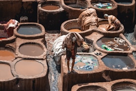 A traditional tannery with several circular stone pits filled with colored liquid can be seen. Workers are engaged in their tasks, handling pieces of leather and fabric. The scene reflects the labor-intensive process of leather tanning, with the earthy tones of the stone contrasting with the vibrant hues of the dye.