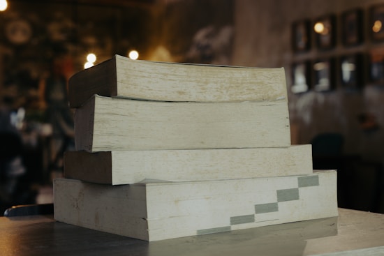 A stack of four large, thick books is placed on a wooden surface. The books have worn, yellowed pages, suggesting age or frequent use. The background is blurred, with a warm tone and small lights, resembling a cozy room or library setting.