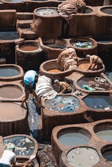 A bustling traditional tannery with a series of round, stone vats filled with various colored liquids for dyeing and treating leather. Workers, some wearing headscarves, are engaged in handling large sheets of hides, moving them between vats. The scene is rich in earthy tones and reflects the manual labor involved in leather production.