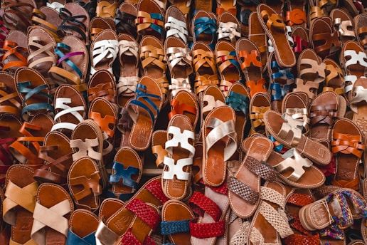 A large collection of sandals is arranged in a visually appealing pattern. The sandals vary in color, style, and texture, displaying craftsmanship and variety. They are primarily leather, with intricate straps and designs.