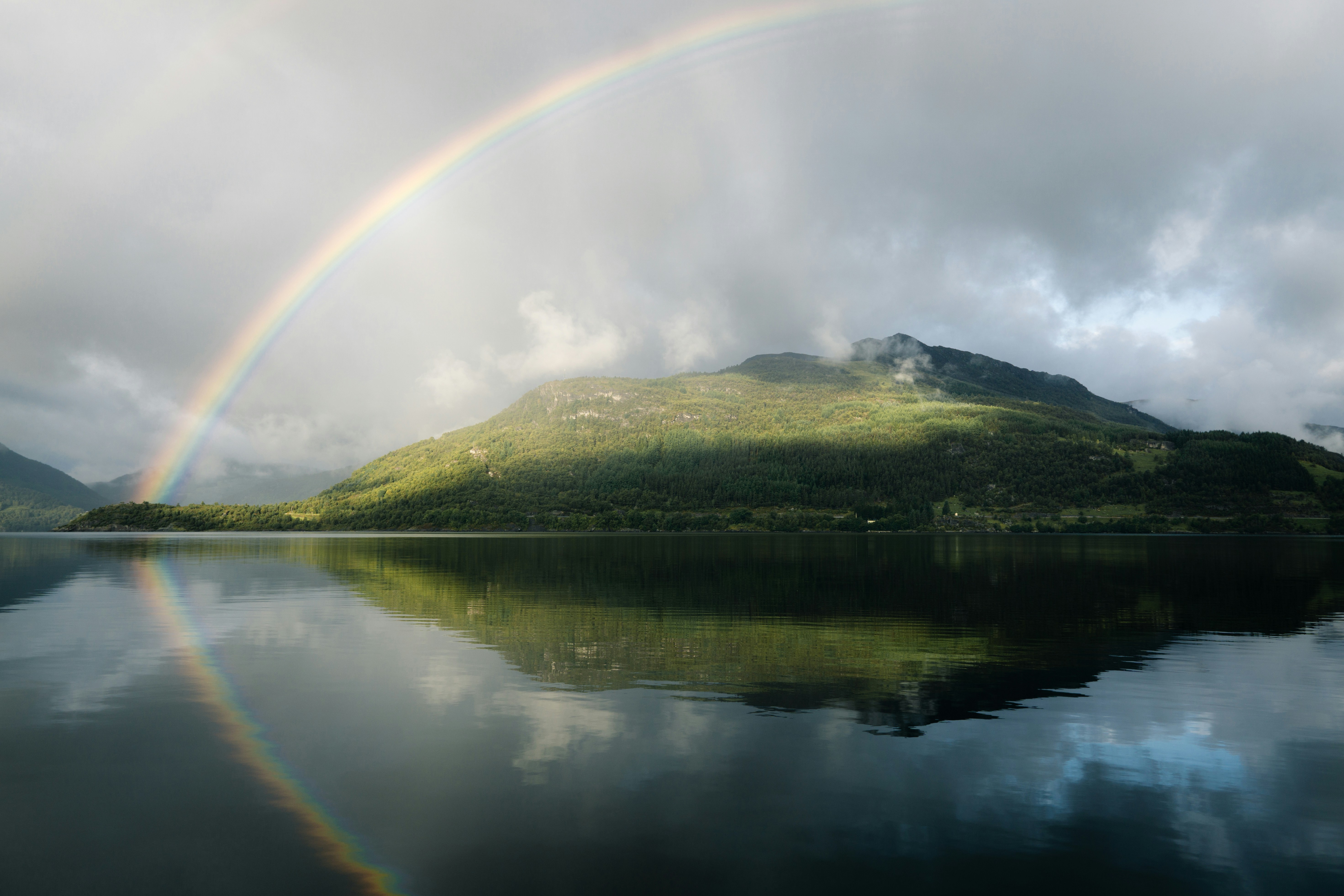 a rainbow over a lake with a mountain in the background, A stunning fjord landscape graced by a vibrant rainbow, adding a touch of enchantment to the majestic natural beauty of the scene