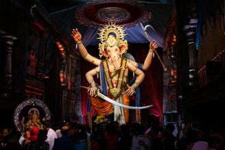 A large, ornate statue of the Hindu deity Ganesha is prominently displayed in a decorated hall. The figure is adorned with a vibrant crown and elaborate jewelry, holding various symbolic objects in its multiple arms. The statue is surrounded by a crowd of people, some of whom are taking photos or praying. The background includes intricate architecture with carvings and draping fabric.