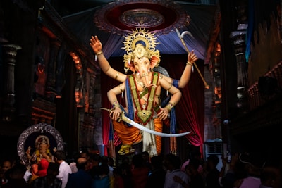 A large, ornate statue of the Hindu deity Ganesha is prominently displayed in a decorated hall. The figure is adorned with a vibrant crown and elaborate jewelry, holding various symbolic objects in its multiple arms. The statue is surrounded by a crowd of people, some of whom are taking photos or praying. The background includes intricate architecture with carvings and draping fabric.