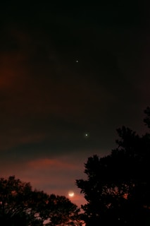 A serene night sky features a crescent moon and two bright celestial objects, possibly planets or stars, against a backdrop of dark clouds. Silhouettes of trees are visible in the foreground, adding depth to the scene.