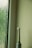 An electric toothbrush stands upright beside a textured glass window with water droplets. The setting appears to be minimalistic, with a light green wall and a faintly lit ambiance.