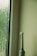 An electric toothbrush stands upright beside a textured glass window with water droplets. The setting appears to be minimalistic, with a light green wall and a faintly lit ambiance.