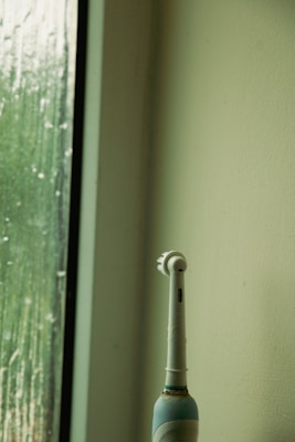 An electric toothbrush stands upright beside a textured glass window with water droplets. The setting appears to be minimalistic, with a light green wall and a faintly lit ambiance.